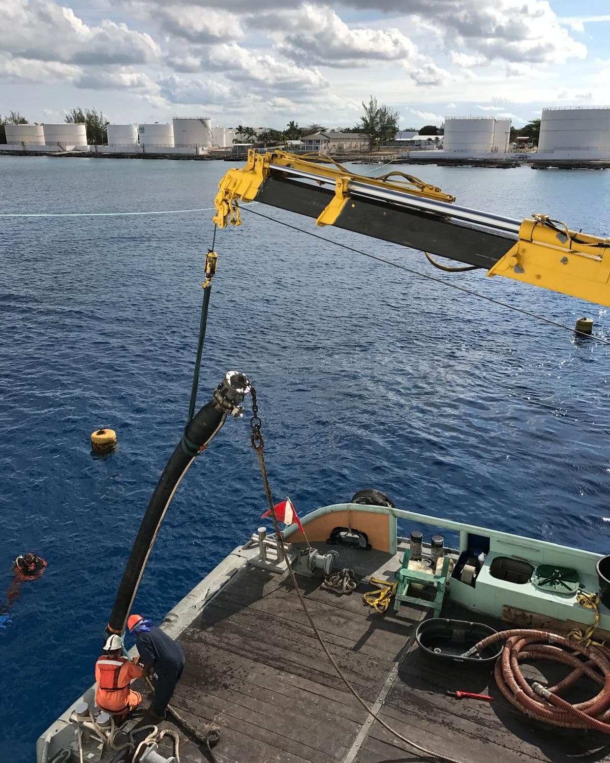 Deckhands in orange safety gear guiding a fuel hose on a yellow crane boom over a workboat stern, shore fuel tanks across the water