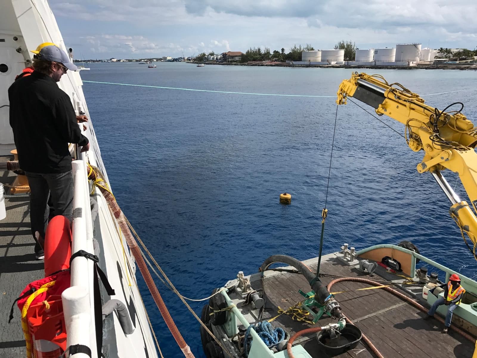 A floating fuel pipeline running across a harbor toward shore storage tanks in the Cayman Islands, a crane workboat alongside
